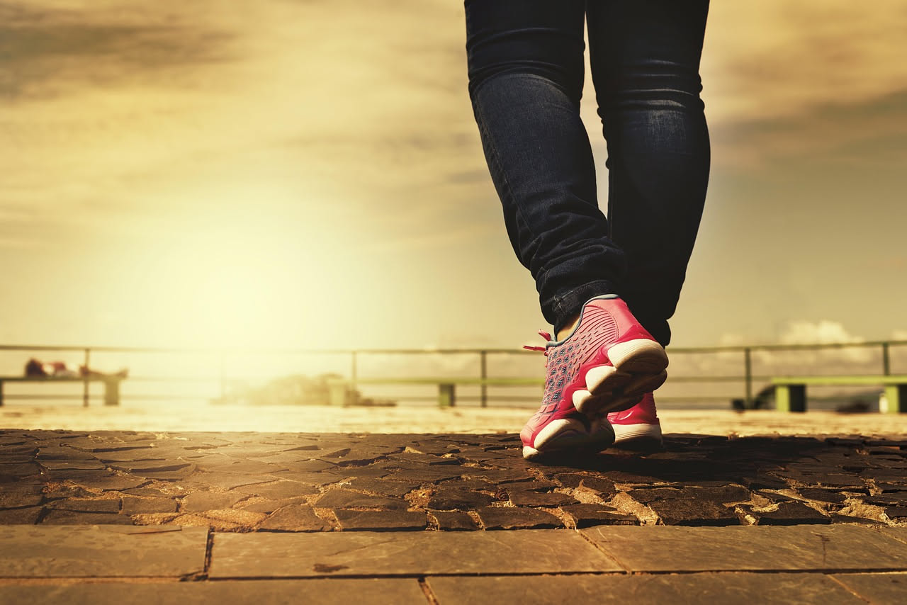 Donna che cammina in un parco verde durante l'alba, indossa abbigliamento sportivo leggero e scarpe da ginnastica, simboleggiando uno stile di vita attivo e salutare