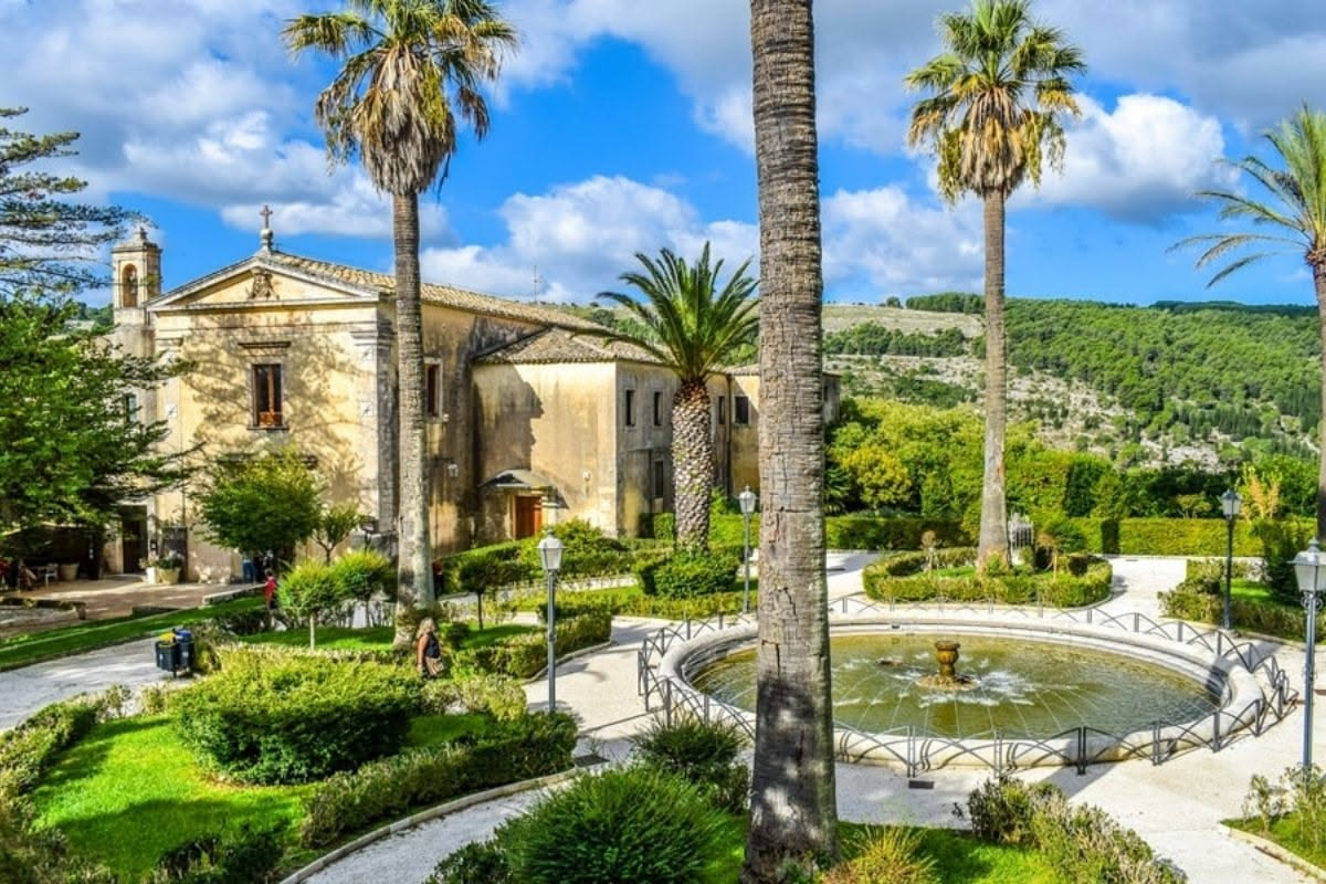 Giardino terrazzato di Villa Hanbury con palme alte, agavi, piante grasse sparse fra sentieri in pietra, mare ligure sullo sfondo al tramonto, vegetazione densa e ordinata