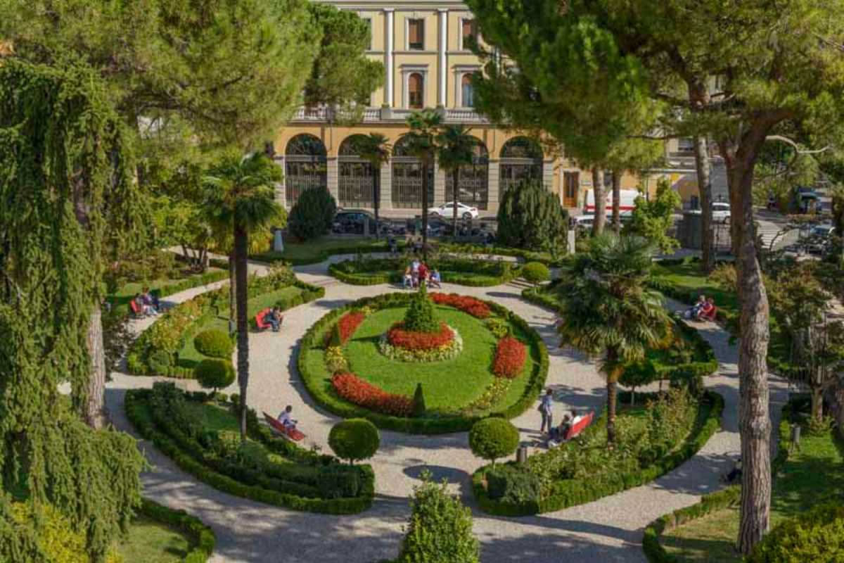 Giardino storico con fontana centrale, alberi ad alto fusto e aiuole regolari circondate da muri in travertino, con palazzo barocco sullo sfondo