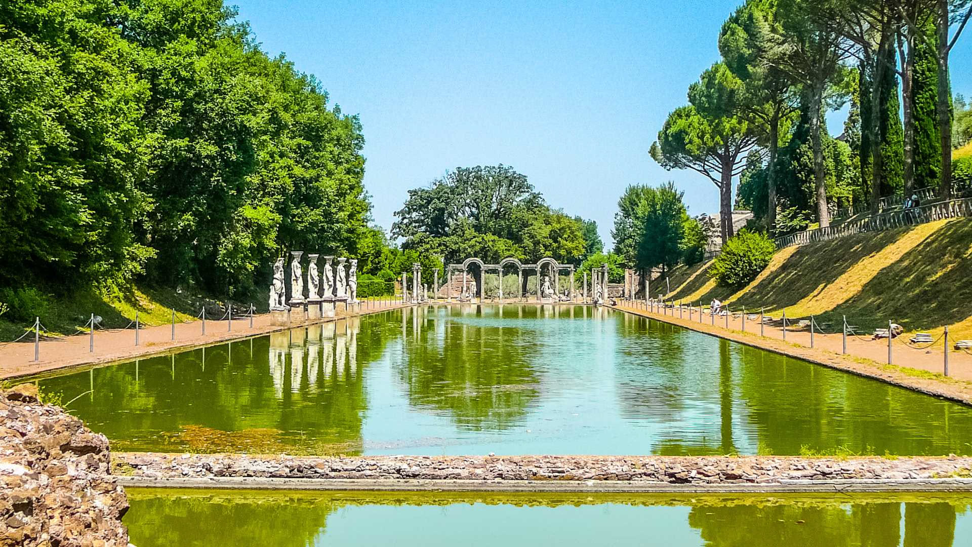 Resti delle terme di Villa Adriana circondati da cipressi scuri, con statue bianche e lo sfondo di oliveti collinari nel tramonto dorato