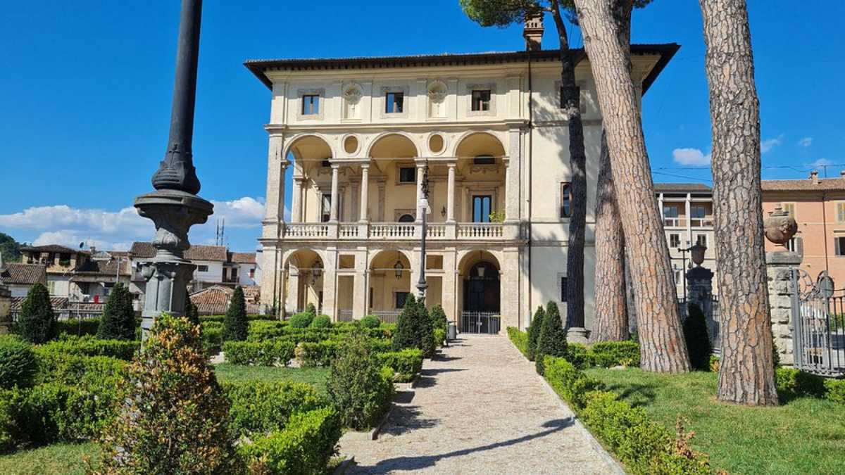 Viale alberato con statue barocche coperte di licheni in un giardino all italiana, alberi ad alto fusto che creano ombre, terracotta rossa di Villa Litta Modignani sullo sfondo