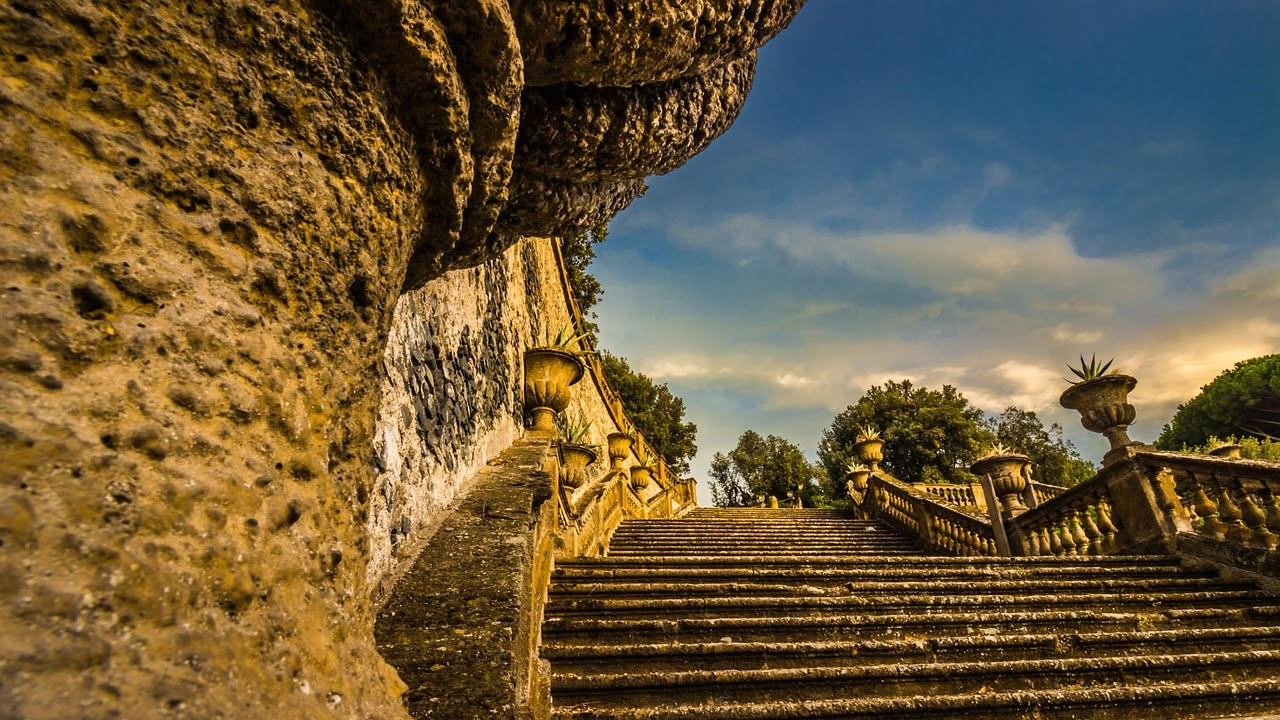 Scala monumentale in pietra che scende nel giardino di Villa Aldobrandini a Frascati, circondata da siepi verdi e fontane, sotto il cielo delle colline laziali