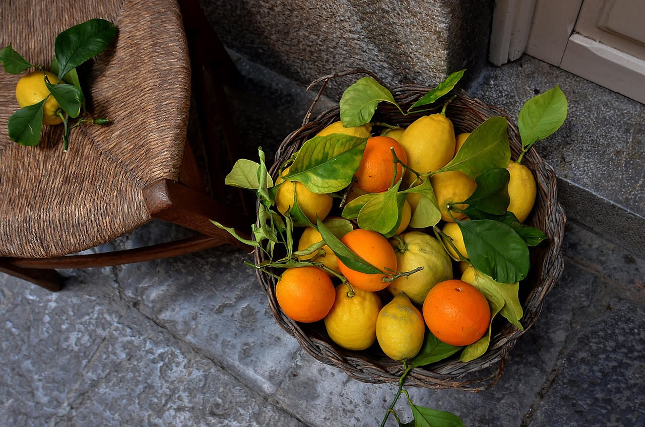 Terrazza di agrumeto siciliano con limoni gialli e foglie verdi rigogliose, borghi di pietra sulle colline sullo sfondo al tramonto, muri a secco e sentieri di terra
