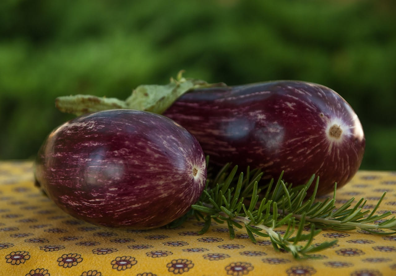 Mani che soppesano una melanzana viola lucida davanti a un banco di melanzane fresche in un mercato estivo, mostrando la cicatrice sul fondo