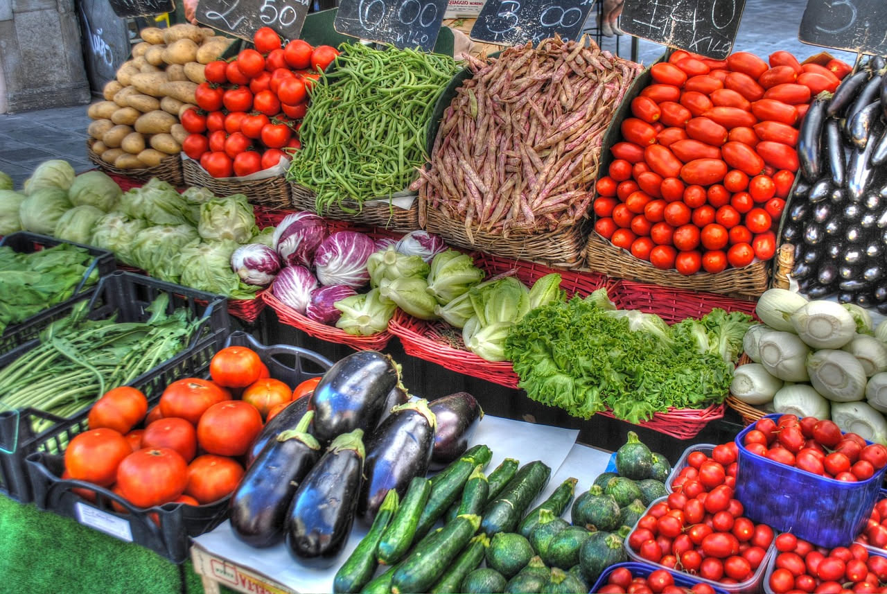 Banco di frutta e verdura al mercato con mele lucide, pomodori e pesche, e le mani di una persona che ne osserva e annusa una da vicino