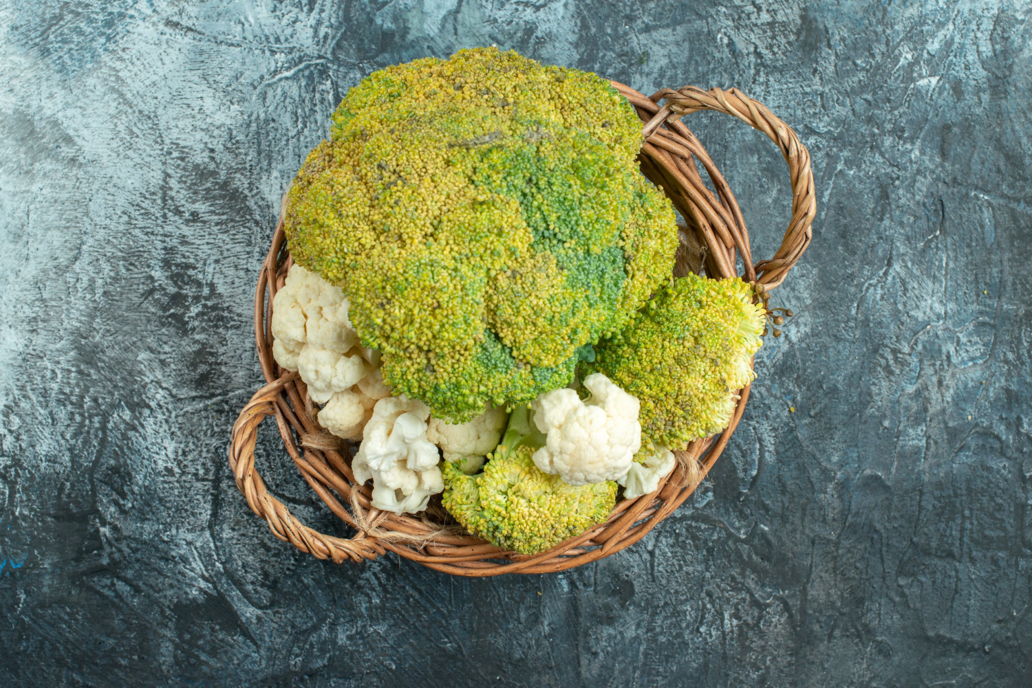 Broccoli con infiorescenze ingiallite e cimette già aperte in un cesto di vimini accanto a cavolfiori